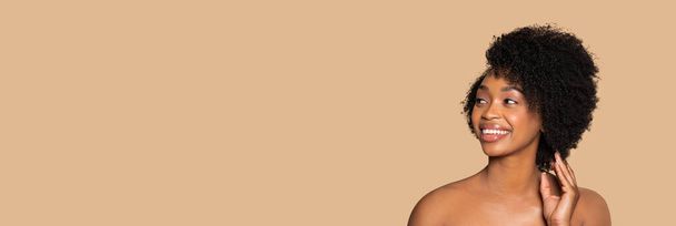 Promienna młoda czarna kobieta z kręconymi, naturalnymi włosami, patrząca na wolną przestrzeń z radosnym uśmiechem, pozująca na beżowym tle, ucieleśniająca naturalne piękno, baner reklamowy - Zdjęcie, obraz
