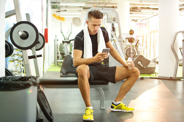 Młody przystojny mężczyzna przy użyciu telefonu podczas wykonywania break w siłowni. - Zdjęcie, obraz