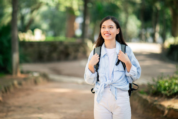Hymyilevä nuori eurooppalainen nainen opiskelija yllään rento vaaleansininen paita ja valkoiset housut solmittu paitahäntä nauttii rauhassa kävellä rauhallinen puistossa reppu, ulkona - Valokuva, kuva