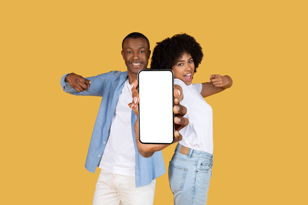 Ενθουσιασμένη χιλιετή ευτυχής African American ζευγάρι παρουσιάζει ένα smartphone με μια κενή οθόνη προς την κάμερα, ιδανικό για την εφαρμογή ή την προώθηση της ιστοσελίδας, σε ένα χαρούμενο κίτρινο φόντο - Φωτογραφία, εικόνα
