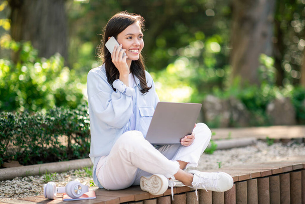Relaxed european young woman student engaged in a phone call while working on a laptop, seated comfortably in a lush park, showcasing multitasking in a tranquil environment - Photo, Image