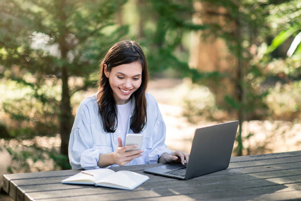 Zufriedene europäische Studentin beim Multitasking mit Smartphone und Laptop an einem Holztisch im Freien, umgeben von üppigem Grün in einer friedlichen Parklandschaft - Foto, Bild