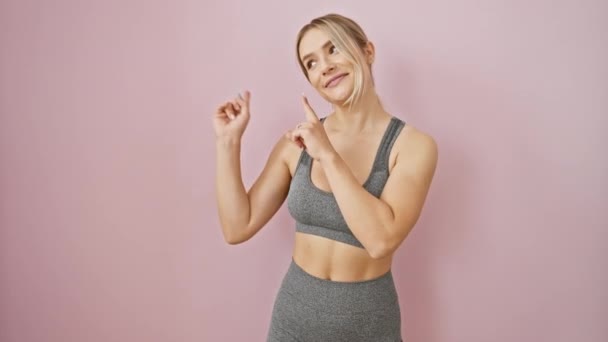 Cheerful young blonde woman confidently sporting activewear, smiling big, pointing to the side; a perfect billboard over isolated pink backdrop. - Footage, Video