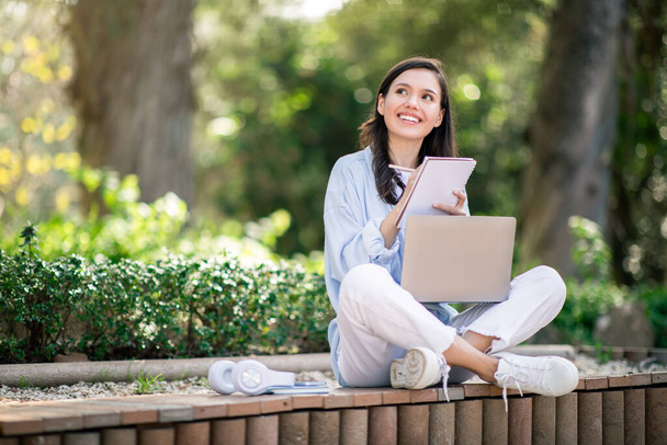 Nachdenkliche junge Studentin mit Laptop, die im Schneidersitz auf einer Parkbank sitzt, ein Notizbuch in der Hand hält und nach oben schaut, möglicherweise Brainstorming oder Planung - Foto, Bild