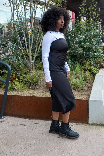 Cette image franche montre une femme élégante portant une longue robe noire sur un haut blanc à manches longues, associée à des bottes de combat noires. Ses cheveux bouclés et naturels encadrent son visage pendant qu'elle traverse une - Photo, image