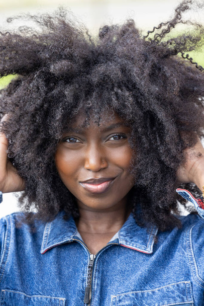 Een close-up portret van een levendige vrouw met een speels afro kapsel, die de camera een subtiele, zelfverzekerde glimlach geeft. Ze draagt een denim jas die een casual maar edgy touch toevoegt aan haar look - Foto, afbeelding