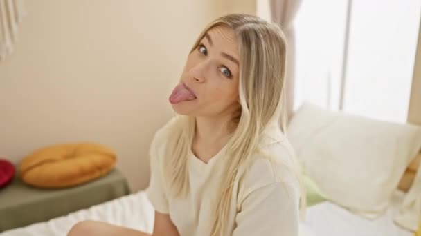 Expression délicieusement amusante sur le visage d'une jeune femme blonde, se tenant joyeusement la langue sur un lit en pyjama, dégageant des vibrations positives dans sa chambre confortable. - Séquence, vidéo