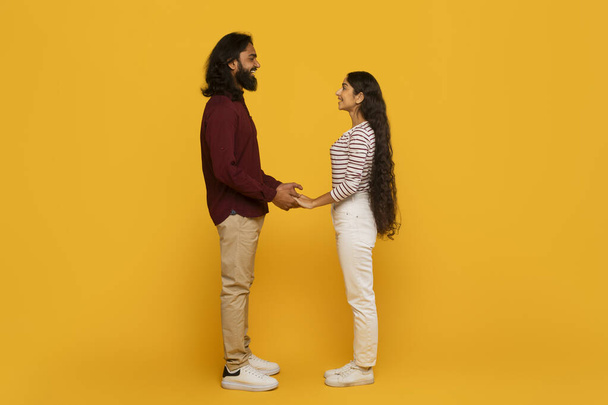 Un hombre y una mujer de pie tomados de la mano, uno frente al otro sobre un fondo amarillo vibrante, retratando una sensación de conexión y unión - Foto, Imagen