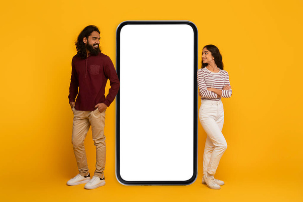 Mężczyzna i kobieta po każdej stronie dużego pustego ekranu smartfona, symulując interakcję na żółtym tle - Zdjęcie, obraz