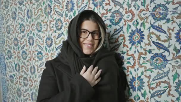 Eine lächelnde Frau mit Brille, in einen Schal gehüllt, posiert vor einem kunstvollen Iznik-Fliesen-Hintergrund in historischer Umgebung.. - Filmmaterial, Video