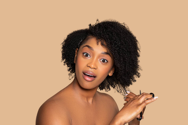 Νεαρή μαύρη γυναίκα με μια έκφραση έκπληξης με ψαλίδι στο χέρι, κόβει τα ογκώδη σγουρά μαλλιά της, σε μαλακό μπεζ φόντο - Φωτογραφία, εικόνα