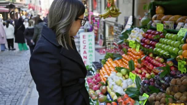 Zamyšlená žena s obchody s brýlemi v pestrobarevném ovocném stánku na rušné istanbulské ulici, odrážející městský život a cestovní ruch. - Záběry, video