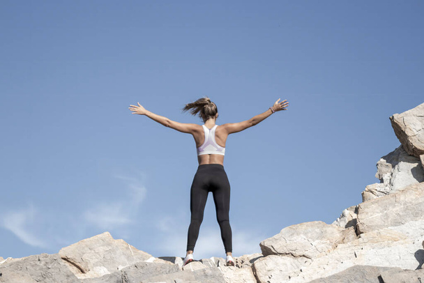 Uma pessoa está triunfalmente no topo de terrenos rochosos, braços estendidos, sob um céu azul claro, incorporando liberdade e realização - Foto, Imagem