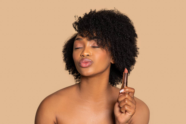 Black woman with curly hair confidently holds lipstick, puckering her lips with playful expression, embodying beauty and fun on beige background - Foto, afbeelding