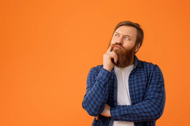 Рудий чоловік у простій сорочці задумливо торкається бороди, думаючи над пропозицією, не звертаючи уваги на сміливе вираження на вільному просторі, на помаранчевому тлі в студії, рекламний банер - Фото, зображення