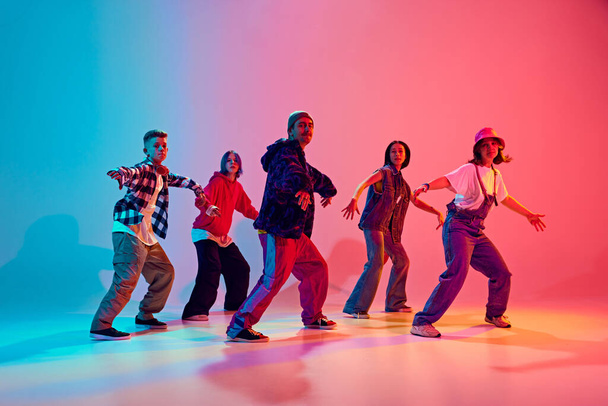 Energetic young dancers in street fashion clothes dance in neon light against gradient colorful studio background. Concept of hobby, sport, fashion and style, action, youth culture, music and dance. - Photo, Image