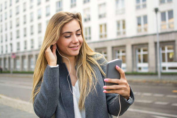 Портрет улыбающейся блондинки со смартфоном, слушающей музыку на городской улице. - Фото, изображение