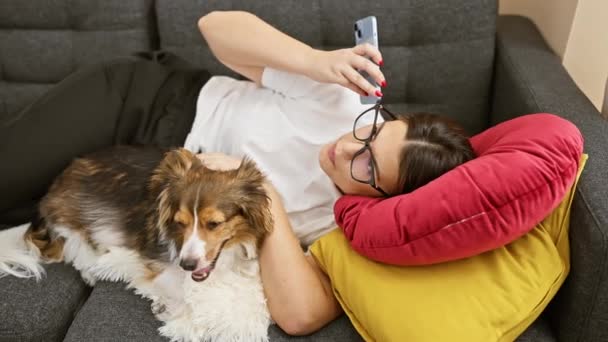 Een ontspannen vrouw in een bril en een hond loungen op een bank met een smartphone binnen - Video