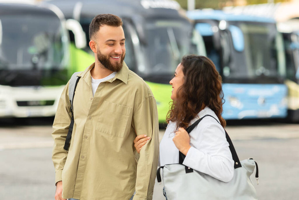 Uśmiechnięta młoda para spacerująca z torbami na dworcu autobusowym i rozmawiająca, szczęśliwy millenium mężczyzna i kobieta cieszący się razem, kochający małżonkowie gotowi do podróży i nowych przygód, wolna przestrzeń - Zdjęcie, obraz