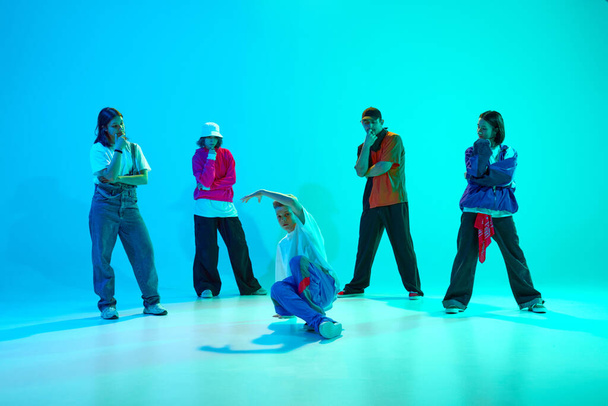 Dance battles. Young guy performing breakdance moves with his dance team in neon light against gradient colorful studio background. Concept of hobby, sport, fashion, youth culture, music and dance. - Zdjęcie, obraz