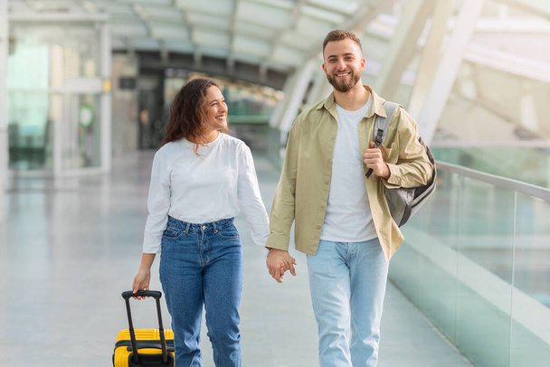 Retrato de pareja joven y romántica caminando juntos en la terminal del aeropuerto, sonriente hombre y mujer milenarios llevando equipaje, tomados de la mano y mirando la cámara, listos para viajar de luna de miel - Foto, imagen