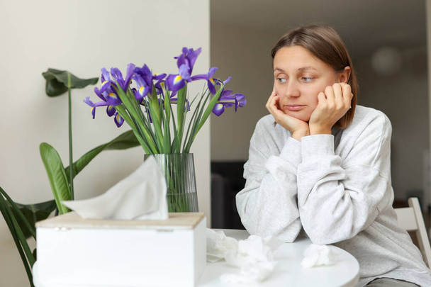 Seasonal allergy. Unhealthy allergic young woman uses paper tissues, suffers from runny nose, sneezes from flowers pollen at kitchen home by table. Girl with flu, itching or cough, allergy rhinitis. - Photo, Image