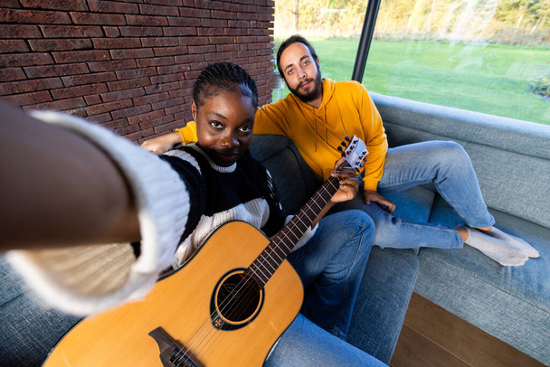 En esta acogedora escena, dos amigos comparten un momento de música y compañía. Una mujer, extendiendo su brazo para una selfie, sostiene una guitarra, mientras que un hombre a su lado se relaja en el sofá. La habitación soleada y - Foto, imagen