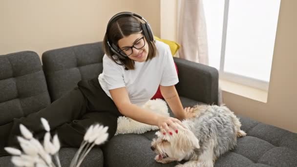 Een jonge vrouw geniet van muziek met koptelefoon, aaien haar hond op een bank thuis. - Video