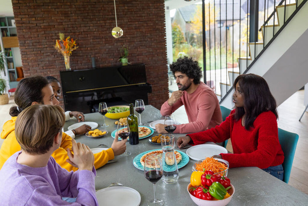 Захоплюючий знімок різноманітної групи друзів, які обговорювали смачний обід у стильній домашній обстановці. Стіл багатий італійськими стравами, а відкрита кімната - Фото, зображення