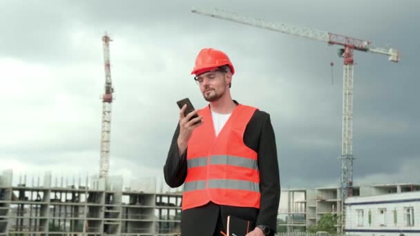 Portret van professionele bouwer in beschermende helm, vest en bril met smartphone en chatten. Langzame beweging - Video