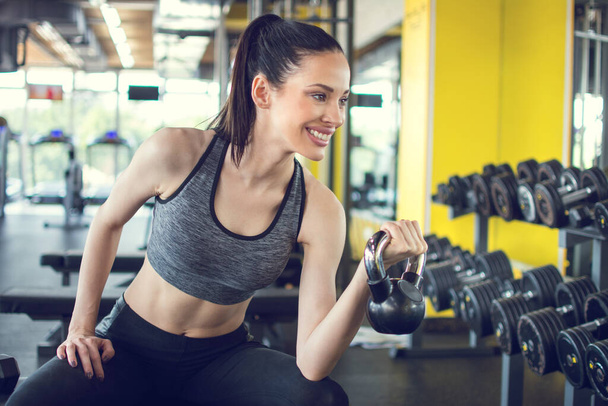 Jeune femme en forme assise sur le banc d'exercice et la cloche de bouilloire de levage dans la salle de gym - Photo, image