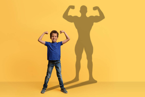 A cheerful boy in a blue shirt flexes his muscles with a big smile, casting a strong superhero silhouette on a yellow background, illustrating youthful confidence and dreams - Photo, Image