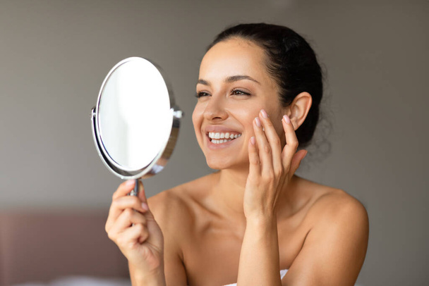 Wesoła młoda kobieta uśmiecha się stosując krem nawilżający trzymając lustro, promując piękno i pielęgnację twarzy, zbliżenie w nowoczesnym wnętrzu sypialni. Pojęcie odmłodzenia dla gładkiej zdrowej skóry - Zdjęcie, obraz