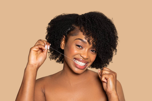 Africká americká žena s kudrnatými vlasy radostně zubní nit, ukazuje široký, poutavý úsměv na teplém béžovém pozadí, podpora zdraví zubů - Fotografie, Obrázek