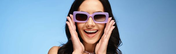 Μια κομψή γυναίκα με μωβ γυαλιά ηλίου, τα χέρια στο πρόσωπο, αποπνέουν εμπιστοσύνη σε μια καλοκαιρινή φωτογράφιση στούντιο σε μπλε φόντο. - Φωτογραφία, εικόνα