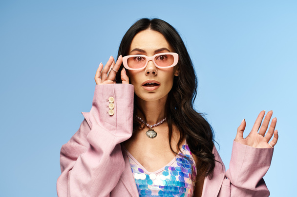A fashionable woman exudes summertime vibes in a pink jacket and sunglasses against a vibrant blue studio backdrop. - Foto, afbeelding