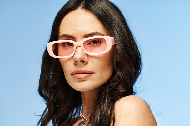 Egy rózsaszín napszemüveges, stílusos nő magabiztosan pózol, nyári rezgéseket áraszt egy stúdióban, kék háttérrel.. - Fotó, kép