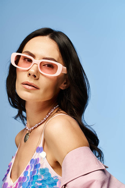 Una mujer elegante con gafas de sol de color rosa y un top rosa posa con confianza sobre un fondo de estudio azul, exudando vibraciones de verano. - Foto, Imagen