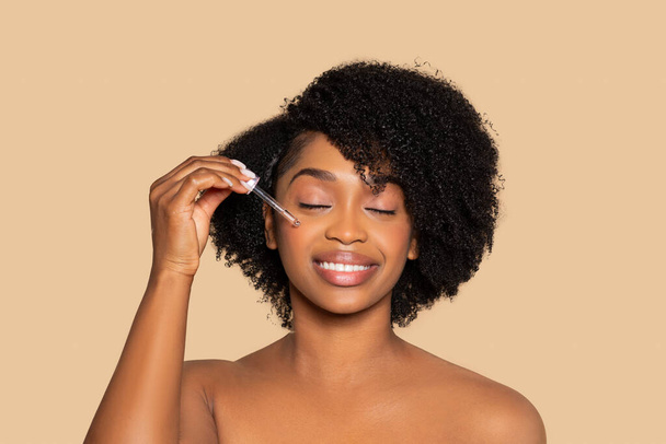 Mujer negra joven satisfecha con los ojos cerrados aplica suavemente suero facial con gotero, disfrutando de su rutina de cuidado de la piel contra el fondo beige - Foto, imagen