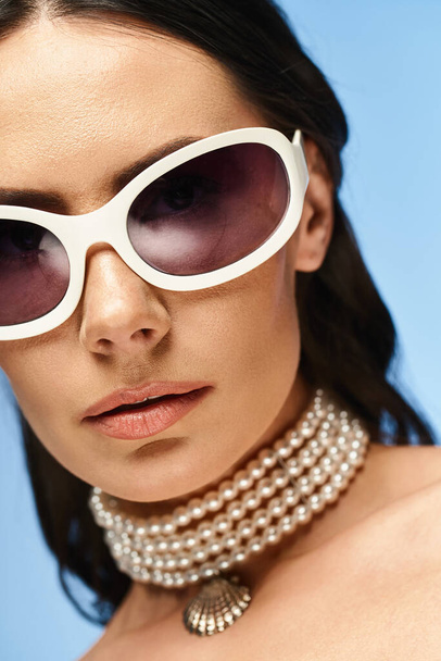 Stylová žena vyzařuje eleganci v slunečních brýlích a perlový náhrdelník na pulzujícím modrém pozadí. - Fotografie, Obrázek