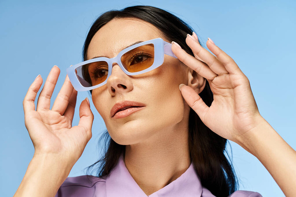 Módní žena se slunečními brýlemi a fialovou košilí pózující sebevědomě ve studiu s modrým pozadím, vyzařující letní vibrace. - Fotografie, Obrázek