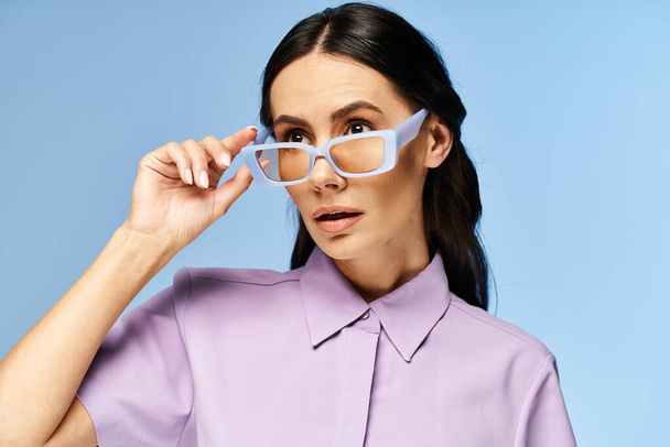 Una mujer elegante con una camisa púrpura y gafas de sol posa con confianza sobre un vibrante fondo de estudio azul, exudando vibraciones veraniegas. - Foto, imagen