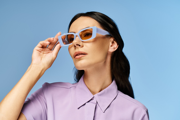 Stylowa kobieta w fioletowej koszuli i okularach przeciwsłonecznych z pewnością pozuje na żywe niebieskie tło w studio. - Zdjęcie, obraz