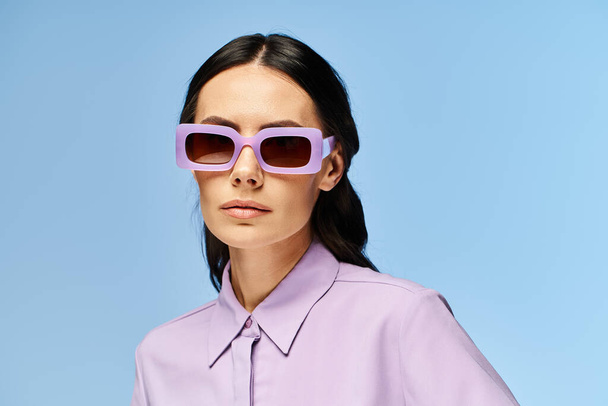 Una mujer de moda está disfrutando de las vibraciones del verano, vistiendo atuendo púrpura y gafas de sol de moda contra un fondo de estudio azul. - Foto, imagen