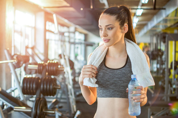 Portrait of beautiful young sporty woman with water bottle and towel at gym. - Photo, Image