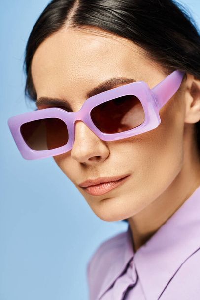 Modna kobieta emanuje letnimi wibracjami w fioletowej koszuli i modnych okularach przeciwsłonecznych na niebieskim tle studia. - Zdjęcie, obraz
