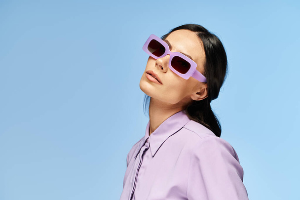 Стильная женщина в фиолетовых солнцезащитных очках и рубашке позирует на синем фоне в летней студии. - Фото, изображение