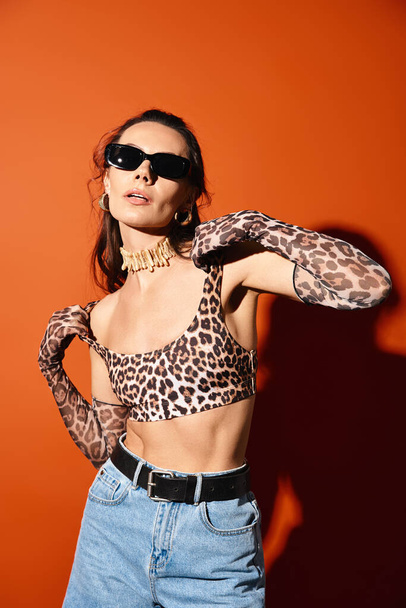 Una mujer de moda en un top de estampado de leopardo y gafas de sol posa con confianza en un entorno de estudio sobre un fondo naranja. - Foto, Imagen