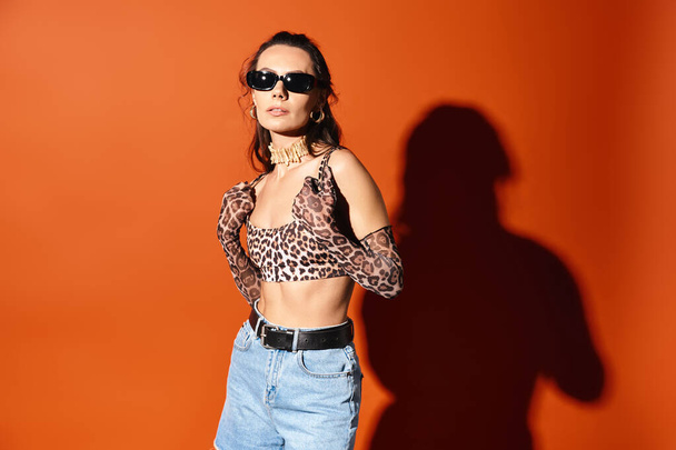 Stylowa kobieta w okularach przeciwsłonecznych pozuje w top z nadrukiem lamparta i jeansowe szorty na pomarańczowym tle, emanujące letnimi wibracjami mody. - Zdjęcie, obraz