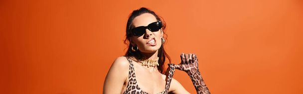 A stylish woman in a leopard print dress poses with a leopard print in a studio setting against an orange background. - Photo, Image